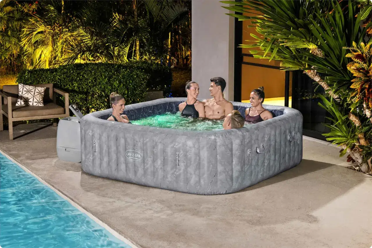 Grupa przyjaciół cieszy się relaksem w jacuzzi ogrodowym Lay-Z-Spa, otoczonym tropikalną roślinnością i basenem.